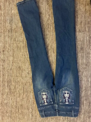 Blå jeans med broderade bakfickor - Säljer ett par blå jeans med snygga broderade detaljer på bakfickorna. Jeansen har klassisk femficksdesign, dragkedja och knapp framtill. Materialet är jeans och passformen är rak med normal midja. Perfekt för dig som gillar unika detaljer på dina jeans.