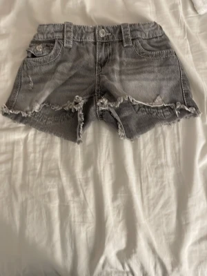 Grå denimshorts med broderade fickor - Säljer ett par grå jeansshorts med fransig kant och snygga broderade detaljer med paljetter på bakfickorna. Shortsen har normal passform, klassiska fickor fram och bak samt hällor för bälte. Perfekta för sommarens chill dagar.