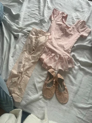 Balletskor i ljusrosa skinn från Bloch och en rosa ballet body med fladder vid midjan, ljus rosa ballet byxor - Säljer ett par klassiska balletskor från Bloch i ljusrosa skinn med rund tå och elastiskt band över foten. Skorna har en mjuk platt sula och en söt rosettdetalj framtill. Perfekta för dans och balettträning. Och med de får du kläderna med på köpet dessa kläder är dock för barn från 8-10 dessa kläder och skor har knappt används ksk 2-3 gånger