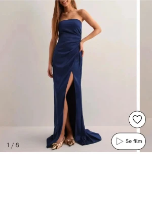 Balklänning - Säljer en marinblå axelbandslös långklänning med slits framtill. Klänningen har en elegant, figurnära passform och är tillverkad i ett glansigt material som ger en lyxig känsla. Perfekt för bal eller festliga tillfällen där du vill sticka ut. Använd 2 gånger!