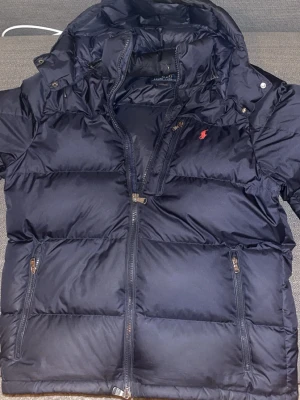 Mörkblå pufferjacka från Polo Ralph Lauren - Snygg mörkblå pufferjacka från Polo Ralph Lauren med röd logga på bröstet. Jackan har flera dragkedjefickor, hög krage och justerbar huva. Perfekt för kalla dagar och har en sportig vibe. Materialet är polyester och jackan är quiltad för extra värme.
