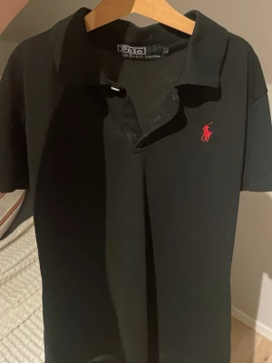 Svart pikétröja från Ralph Lauren - Klassisk svart pikétröja från Ralph Lauren med röd broderad logga på bröstet. Tröjan har krage, korta ärmar och knäppning framtill. Materialet är mjuk bomull som känns skönt mot huden. Perfekt för en stilren och enkel look. Storlek L i barn. Pris kan förhandlas