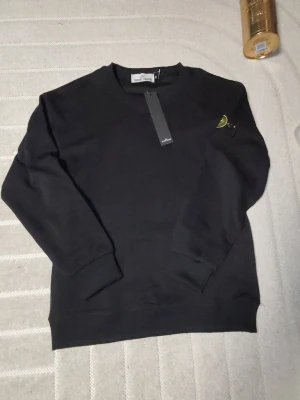 Svart sweatshirt från Stone Island - Svart sweatshirt från Stone Island med klassisk rund hals och ribbade muddar. Tröjan har den ikoniska Stone Island-loggan på vänster ärm, fäst med två knappar. Helt ny Stone Island tröja i storlek M. 👕Det som ingår i priset är endast tröjan. 🎨⬛:Färg Svart