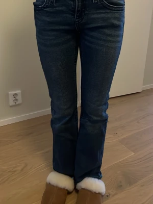 Mörkblå bootcut jeans med detaljer - Säljer ett par mörkblå jeans med bootcut-snitt och snygga detaljer på bakfickorna. Jeansen har klassisk femficksdesign, silverfärgade knappar och är tillverkade i jeansmaterial med lätt tvättad look. Perfekt för dig som gillar en avslappnad men stilren vibe.