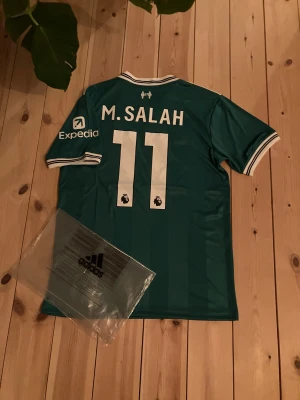 M. Salah #11 Liverpool tröja - Bilderna visar en grön Liverpool-tröja från Adidas med M. Salah och nummer 11 på ryggen, samt tydliga Adidas-taggar och loggor. Ingen sko finns med på bilderna. Inte riktig men jättebra kvalite