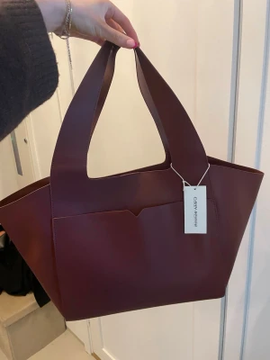 Brun axelväska från Carin Wester - Säljer en stilren brun axelväska från Carin Wester i slätt skinn. Väskan har breda handtag, en yttre ficka och rymligt huvudfack. Perfekt för dig som gillar minimalistisk design och vill ha plats för allt du behöver. Snygg att bära över axeln och passar till många olika outfits.