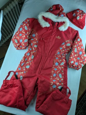 Vintage röd blommig vinteroverall och mössa från European Kids Lindex - Vintage röd vinteroverall från European Kids Lindex med blommigt mönster i blått och gult på ärmar och sidor. Huva med vit fuskpäls, dragkedja framtill och elastiska muddar. Overallen har praktiska fotremmar och matchande röd mössa med ull. Perfekt för kalla dagar! The elastic around the ankles have lost their stretch. I can't do up the zip the last 2cm. There are signs of age such as some fraying.
