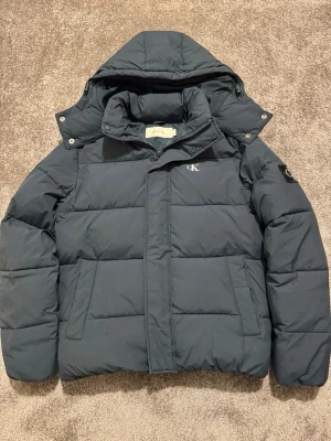 Calvin Klein Jacka - Säljer en mörkblå pufferjacka från Calvin Klein Jeans Skicket är 10/10 och knappt använd, har e-kvitto som kan skickas vid köpet. Nypris: 2400kr. Pris är för mesta dels prutat o klart. Mått: 45cm Axelbredd, 60cm längd. 