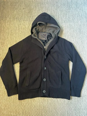 Svart hoodie-kofta från GANT, L - Svart hoodie-kofta från GANT i storlek L. Tröjan har både dragkedja och knappar framtill, huva med grått foder och två fickor. Materialet är en mix av ull och bomull med polyesterfoder i huvan. Ribbstickade muddar vid ärmslut och nederkant ger en schysst look. Den har en defekt vid ärmen som man ser