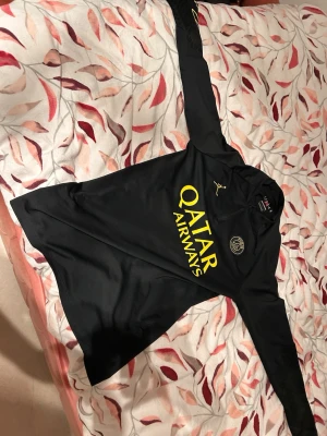 Svart PSG träningsjacka Jordan - Svart långärmad träningsjacka från Jordan x PSG med gul Qatar Airways-tryck på bröstet och PSG-logga. Jackan har halv dragkedja och är gjord i ett lätt syntetmaterial som passar perfekt till fotbollsträning. Snygg och sportig design med gul detalj på ärmen.