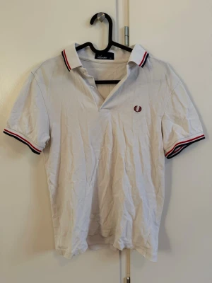 Vit piké från Fred Perry - Klassisk vit pikétröja från Fred Perry med röd och marinblå rand på krage och ärmslut. Ikonisk Fred Perry-logga broderad på bröstet. Tillverkad i bomull och har korta ärmar samt normal passform. Perfekt till jeans eller shorts.