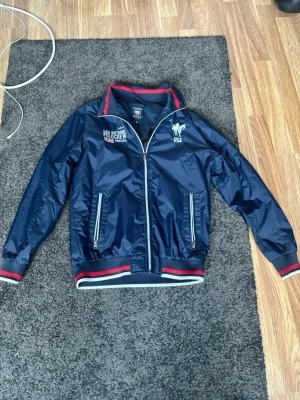 Mörkblå vindjacka från Polo Club - Snygg mörkblå vindjacka från Polo Club med broderad logga och text på bröstet. Jackan har dragkedja framtill, två fickor med dragkedja och ribbade muddar med röd och vit rand. Perfekt för dig som gillar sportig stil och vill sticka ut.