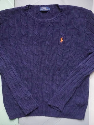 Marinblå stickad tröja Polo Ralph Lauren - Säljer en marinblå stickad tröja från Polo Ralph Lauren i storlek M. Tröjan har ett klassiskt kabelstickat mönster och en rund hals. På bröstet finns den ikoniska orangea Polo-loggan broderad. Perfekt för dig som gillar stilrena och tidlösa plagg.