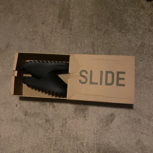 Adidas Yeezy Slide Onyx strl 44 1/2 - Säljer ett par svarta Adidas Yeezy Slide Onyx i storlek 44 1/2. Slidesen har en chunky, räfflad sula och är tillverkade i syntetmaterial. Minimalistisk design med öppen tå och platt sula, perfekt för chill dagar. Box och etikett medföljer.