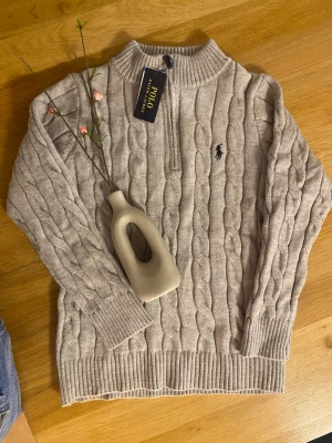 Beige stickad half zip från Polo Ralph Lauren - Säljer en grå stickad tröja från Polo Ralph Lauren med half zip och grov kabelstickning. Tröjan har ribbade muddar, hög krage och den klassiska broderade loggan på bröstet. Perfekt för dig som gillar stilren och tidlös design.