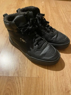 helt nya svarta höga sneakers från Puma - Säljer ett par svarta höga sneakers från Puma med snörning och mjukt foder på insidan. Skorna har en rund tå, platt sula och är tillverkade i skinnliknande material. Perfekta för dig som gillar sportig stil och vill ha extra värme runt ankeln. 