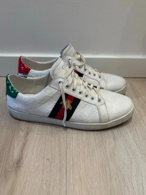 Gucci vita sneakers  - Gucci sneakers i vitt skinn med broderad bi på sidan och klassiska ränder i rött och grönt. Baksidan har metallicröd och metallicgrön häl med ormskinnsmönster. Skorna har snörning och rund tå, samt präglat GG-mönster över hela skon. Kom dm för eventuella frågor 🧐 pris kan diskuteras 💯Om man tvättar dem ser de helt nya ut och i perfekt skick  😉🤩