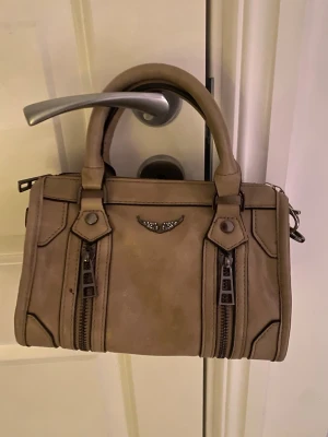 Beige handväska med dragkedjor - Säljer en beige handväska i skinnimitation med två rejäla handtag och snygga svarta detaljer. Väskan har dekorativa dragkedjor framtill och ett litet emblem i silver. Perfekt storlek för det viktigaste och har en cool, boxig form.