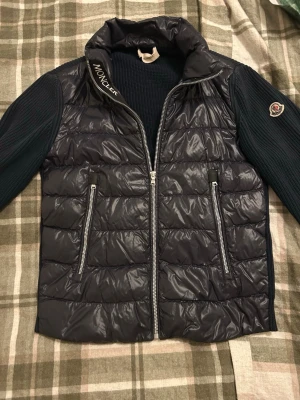 Mörkblå cardigan från Moncler - Säljer en mörkblå pufferjacka från Moncler med stickade ärmar och quiltad front. Jackan har två dragkedjeförsedda fickor och en hög krage med Moncler-logga. Materialmix av polyester och stickad textil, perfekt för dig som vill ha en snygg och sportig look. Nypris: 4700