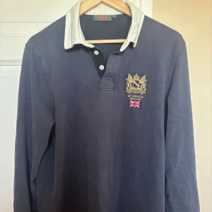 Mörkblå henley från Morris, storlek L - Säljer en mörkblå henley från Morris med långärmad design och kontrasterande ljus krage. Skjortan har broderad logga och brittisk flagga på bröstet samt knappar vid halsen. Materialet är mjuk bomull och kragen har Morris-broderi på baksidan.
