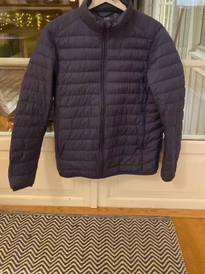 Mörkblå quiltad pufferjacka - Säljer en mörkblå pufferjacka från Uniqlo med quiltad design och dragkedja framtill. Jackan har hög krage och långa ärmar, perfekt för kyliga dagar. Enkel och stilren look som funkar till många outfits.