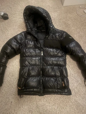 Svart pufferjacka från Alessandro Zavetti - Säljer en svart pufferjacka från Alessandro Zavetti med glansig finish och huva. Jackan har dragkedja framtill, två sidofickor med dragkedja och ett snyggt emblem på ärmen. Insidan har ett mönstrat foder med logotyptryck. Perfekt för kalla dagar. Ett litet hål på baksidan