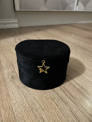 Svart smyckesask med spegel och stjärndetalj - Snygg rund smyckesask i svart sammet med en guldig stjärnberlock framtill. Insidan är ljus och har en liten spegel i locket samt fack för ringar och smycken. Perfekt för att hålla koll på dina favoritsmycken hemma eller på resan.