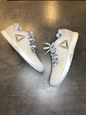 Reebok Crossfit sneakers  - Säljer ett par Reebok Crossfit sneakers med Kevlar-detaljer. Skorna är vita med inslag av ljusgrå och svart mesh, har snörning och platt sula. Reebok-loggan syns tydligt på sidan och hälen. Perfekta för träning och har en sportig look.