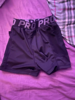 Svarta PRO träningsshorts - Säljer ett par svarta träningsshorts från PRO med bred elastisk midja där det står PRO i grått. Shortsen är tighta och gjorda i syntetmaterial som andas, perfekta för gymmet eller löpning. Snygg och sportig look!