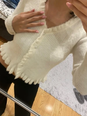 Vit stickad kofta med volangkant - Superfin vit stickad kofta med v-ringning och pärlknappar. Koftan har långärmade ärmar och en croppad passform. Volangdetaljer längs kanten och vid halsen ger en extra söt touch. Perfekt att styla med jeans eller kjol för en trendig look.