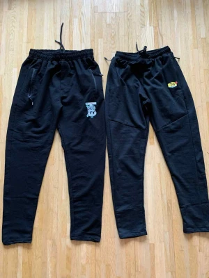 Svarta mjukisbyxor med tryck - Två par svarta mjukisbyxor med elastisk midja och snörning. Ena paret har blått tryck med bokstäver och dragkedjor vid fickorna, det andra har Tn Air-logga i gult och rött. Båda är tillverkade i mjukt bomullsmaterial och har en loose passform som är skön att chilla i.