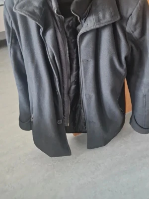 Svart rock med krage och innerfoder - Snygg svart rock med klassisk krage och innerfoder. Jackan har knappar framtill och en stilren design som passar perfekt till kyliga dagar. Materialet känns mjukt och värmande, och den har långa ärmar för extra komfort.