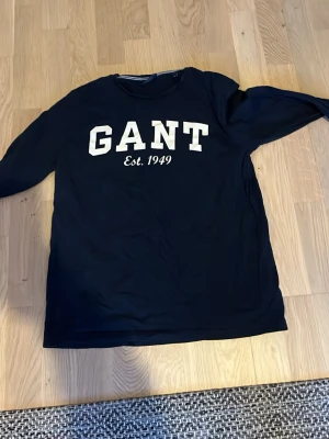Mörkblå långärmad tröja från GANT - Snygg mörkblå långärmad tröja från GANT i storlek M. Tröjan har ett stort vitt tryck med GANT och Est. 1949 på bröstet. Rund hals med diskret randig kant på insidan. Perfekt för dig som gillar klassisk och sportig stil.
