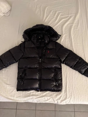 Svart pufferjacka från Polo Ralph Lauren - Svart pufferjacka från Polo Ralph Lauren med glansig finish och röd broderad logga på bröstet. Jackan har huva, två sidofickor med dragkedja och kraftig frontzip. Perfekt för kalla dagar och har en modern, boxig passform.