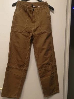 Bruna cargopants med raka ben - Säljer ett par bruna cargopants med raka ben och synliga sömmar. Byxorna har stora fickor fram och bak, bälteshällor och knappstängning. Materialet är slitstark bomull och färgen är en varm brun ton. Perfekt för dig som gillar utility-vibe och streetstyle.