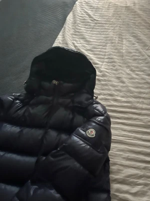 Mörkblå Moncler pufferjacka med huva - Säljer en mörkblå  pufferjacka från Moncler med huva och två sidofickor med dragkedja. Jackan har ett glansigt yttermaterial och Moncler-logga på ärmen. Perfekt för kalla dagar och har en snygg, puffig look som sticker ut.