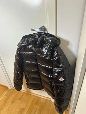 Moncler Maya - Säljer en svart, glansig pufferjacka från Moncler med huva och dragkedja framtill. Jackan har Moncler-logga på ärmen, två sidofickor med dragkedja och en cool, oversized look. Perfekt för dig som vill ha en varm och snygg vinterjacka med streetkänsla.