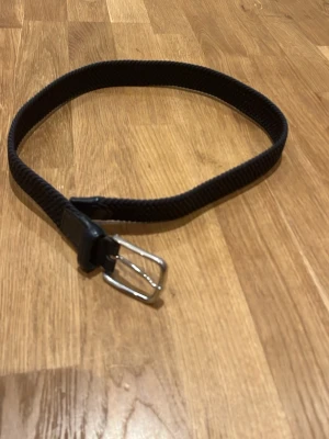 Blått flätad bälte från Lexington - Säljer ett svart flätat bälte med metallspänne och detaljer i skinn. Bältet har en liten röd etikett med vitt kors, inspirerat av danska flaggan. Perfekt för att ge din outfit en snygg touch.