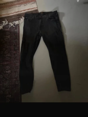 Svarta jeans från Lee, tapered fit - Svarta jeans från Lee med klassisk femficksdesign och knapp med Lee-logga. Modellen är tapered fit med dragkedja i gylfen. Jeansen är tillverkade i bomull och har en mörk tvätt som ger en cool vibe. Perfekt för dig som gillar stilrena och tidlösa jeans.