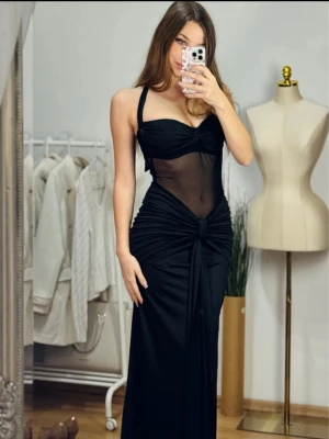 Elegant långklänning med mesh och knyt - Supersnygg svart långklänning med meshdetaljer över magen och ryggen. Klänningen har knyt i nacken och en markerad byst, samt drapering och knut framtill. Perfekt för dig som vill sticka ut med en elegant och trendig look. S / 36 / 8