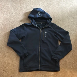 Mörkblå hoodie från Sail Racing - Mörkblå hoodie från Sail Racing med dragkedja hela vägen, huva och diskret logga på bröstet. Tröjan har lång ärm och är tillverkad i ett mjukt material med fleece på insidan. Perfekt för dig som gillar stilren och sportig look.