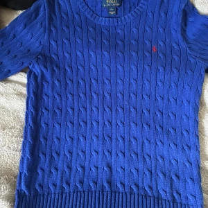 Blå kabelstickad tröja Polo Ralph Lauren - Kabelstickad blå tröja från Polo Ralph Lauren med rund halsringning och lång ärm. Klassisk design med röd broderad logga på bröstet och ribbade muddar. perfekt för dig som är i tonåren och vill ha en snygg märkes tröja 