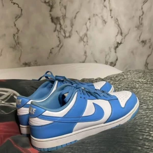 Nike Dunk UNC Low - Nike Dunk Low sneakers i fräsch blå och vit färg. Skorna har klassisk snörning, perforerad tå och platt sula. Swoosh-loggan syns tydligt på sidan och Nike-broderi på hälen. Tillverkade i skinn med syntetdetaljer och har en rund tå för skön passform. Äkta Nike dunk unc. Pris ej hugget i sten 