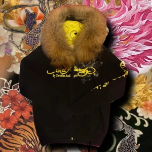 Ed Hardy svart pälsjacka med tryck - Svart jacka från Ed Hardy med stor brun päls runt huvan och färgstarkt tattoo-inspirerat tryck på ryggen. Framsidan har gul logga och detaljer, samt dragkedja. Ärmarna är långärmade med gul text. Jackan har en loose passform och cool streetstil. Står XL men sitter mer som en M/L 