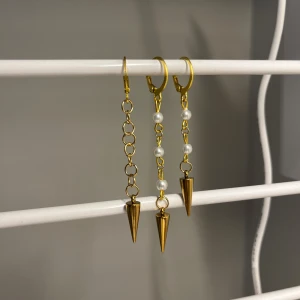 Guldiga örhängen med pärlor och spikes - Unika guldiga örhängen med kedjor och spetsiga spikes i metall. Två av örhängena har vita pärlor längs kedjan för extra detalj, medan det tredje har en enkel kedja. Perfekt för dig som gillar edgy och trendiga smycken. I rostfrittstål och handgjort 