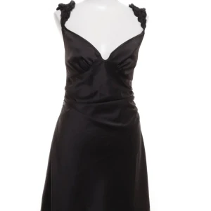 Svart klänning från Adoore - Supersnygg svart klänning från Adoore med v-ringad front och unika axelband med volang  detalj. Klänningen är ärmlös och har en elegant, figurnära passform. Materialet är mjukt och glansigt i polyester med lite stretch för extra komfort. Storlek 38 