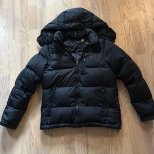 Svart pufferjacka från Polo Ralph Lauren - Svart pufferjacka från Polo Ralph Lauren med huva och dragkedja framtill. Jackan har två fickor med dragkedja och klassisk röd logga på bröstet. Perfekt för kalla dagar och riktigt snygg streetstil.