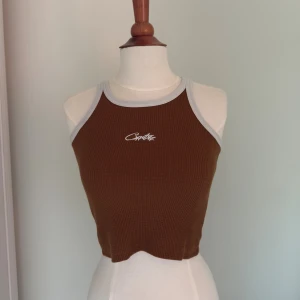Brun croppad linnetopp med smutsig kontrastkant - Säljer en brun croppad linnetopp från Corteiz med ljusgrå kontrastkant runt hals och ärmhål. Toppen är ribbad och har ett broderat vitt Corteiz-logo framtill. Perfekt för en chill och trendig look.