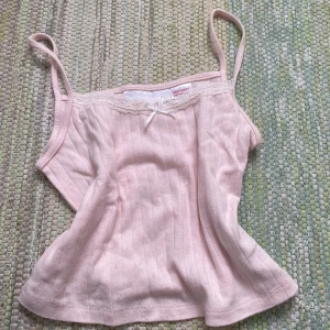 Ljusrosa ribbad linnetopp Gina Tricot - Supersöt ljusrosa linnetopp från Gina Tricot med smala axelband, ribbad struktur och små hålmönster. Toppen har spetskant och en liten rosett framtill för extra gullig touch. Perfekt för varma dagar eller som layering-piece.