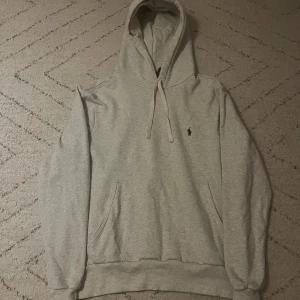 Ljusgrå hoodie från Polo Ralph Lauren - Säljer en stilren ljusgrå hoodie från Polo Ralph Lauren med klassisk broderad logga på bröstet. Tröjan har huva med snörning, känguruficka och ribbade muddar. Perfekt för en chill och avslappnad look. Ej använd! Fraktar inom 1-2 dagar!